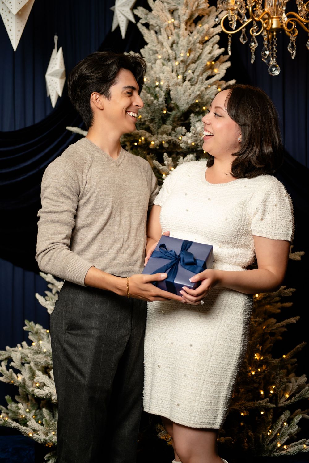 Smiling couple in a festive studio hold a navy gift box tied with a blue ribbon beside a flocked Christmas tree and chandelier.