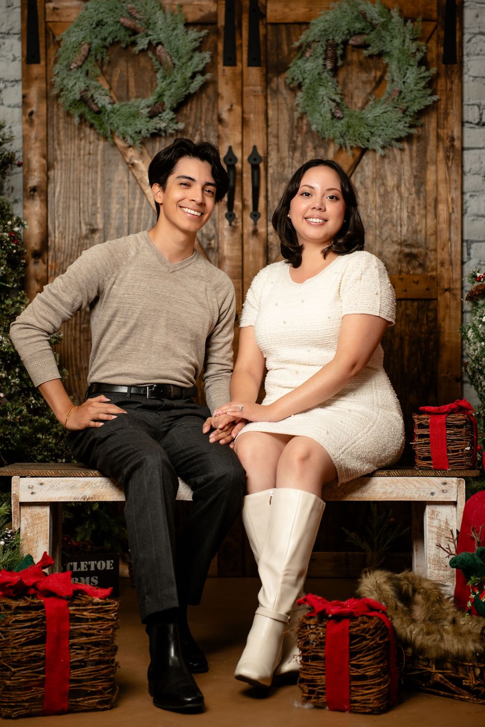 Smiling couple sits holding hands on a wooden bench with red-ribboned gift baskets in front of rustic wreath-decorated doors.