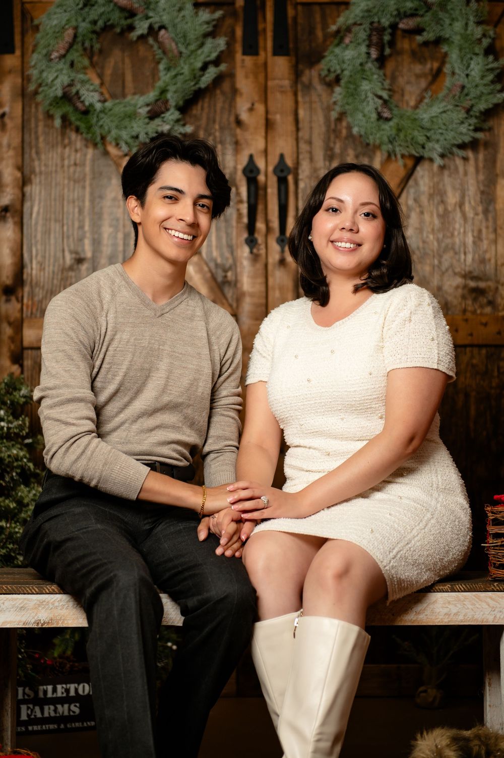 Smiling couple sitting close together holding hands on a wooden bench in front of a rustic barn door with evergreen wreaths.