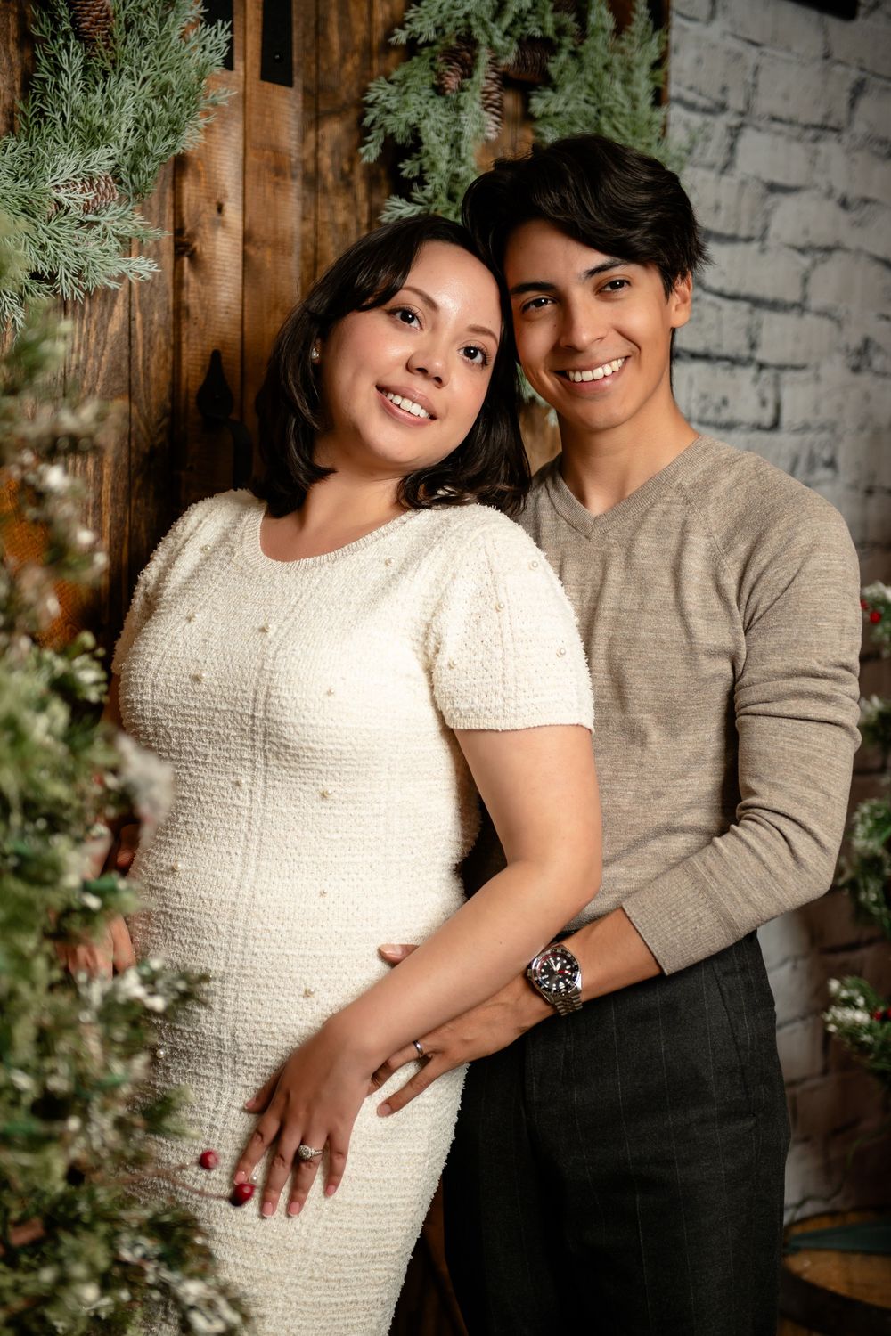 Smiling couple embracing in a cozy holiday photoshoot, she in a white knit dress and he in a beige sweater, framed by greenery.