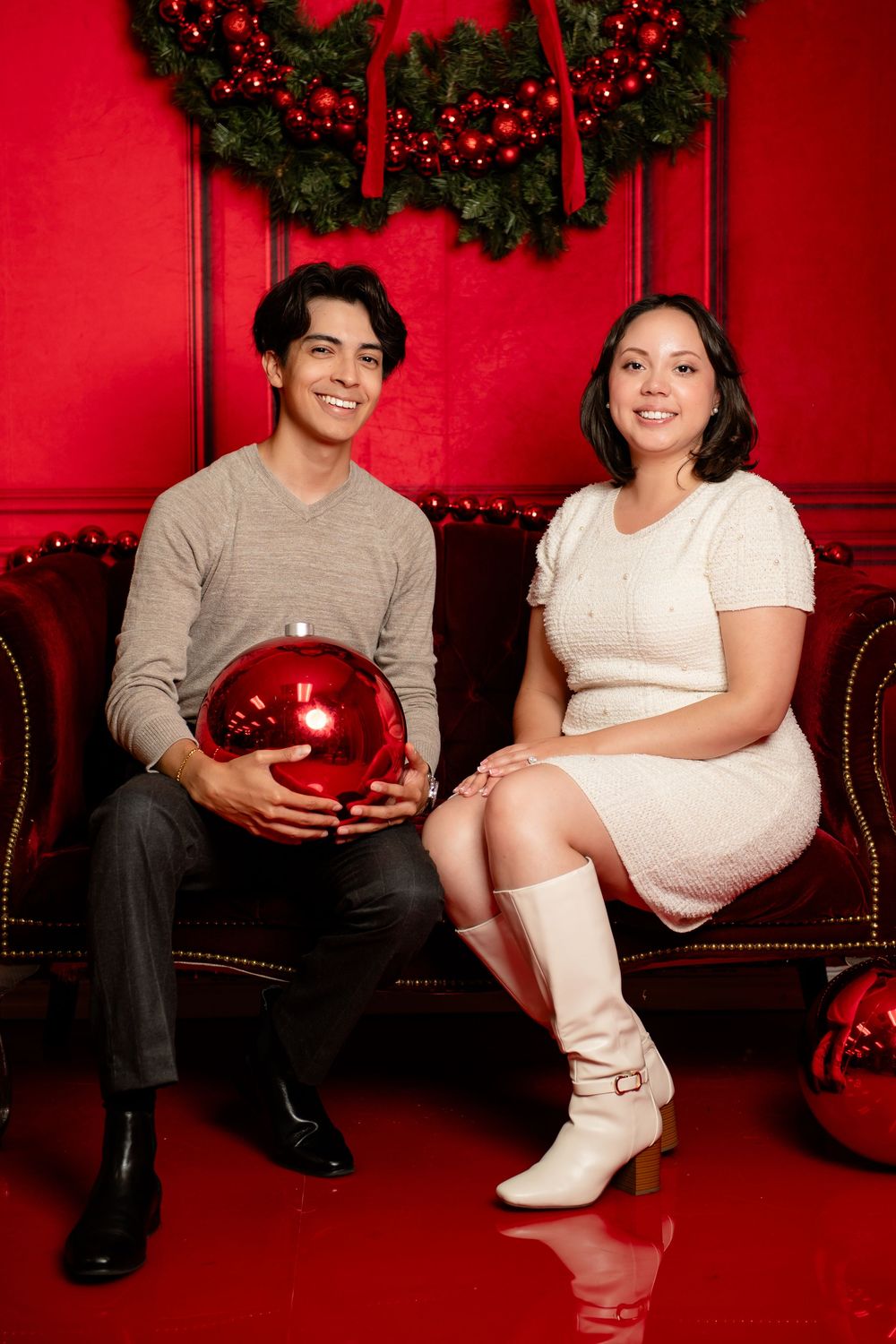 Smiling man and woman sit on a red velvet sofa holding a shiny red ornament beside a festive Christmas wreath.