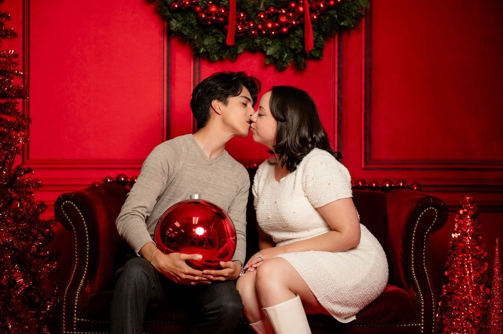 Couple sharing a kiss on a red velvet sofa while holding a large red ornament, with a festive wreath and red holiday backdrop.