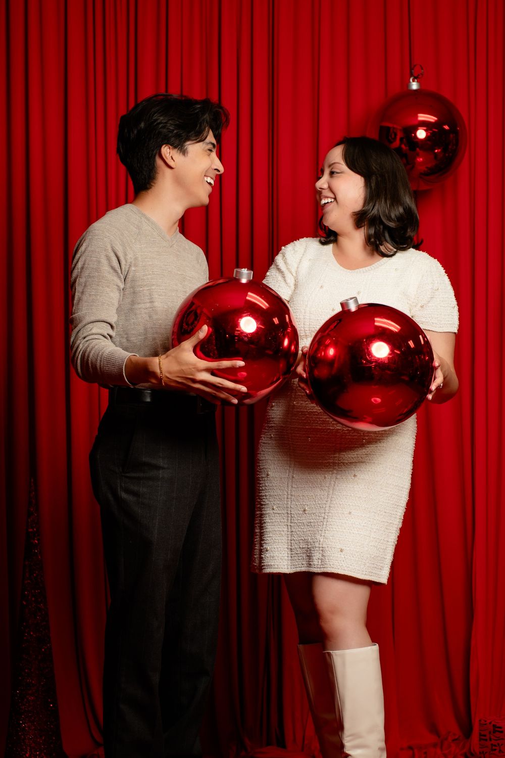 Smiling couple laughing while holding giant red Christmas ornaments against a red velvet curtain in a festive holiday photoshoot.