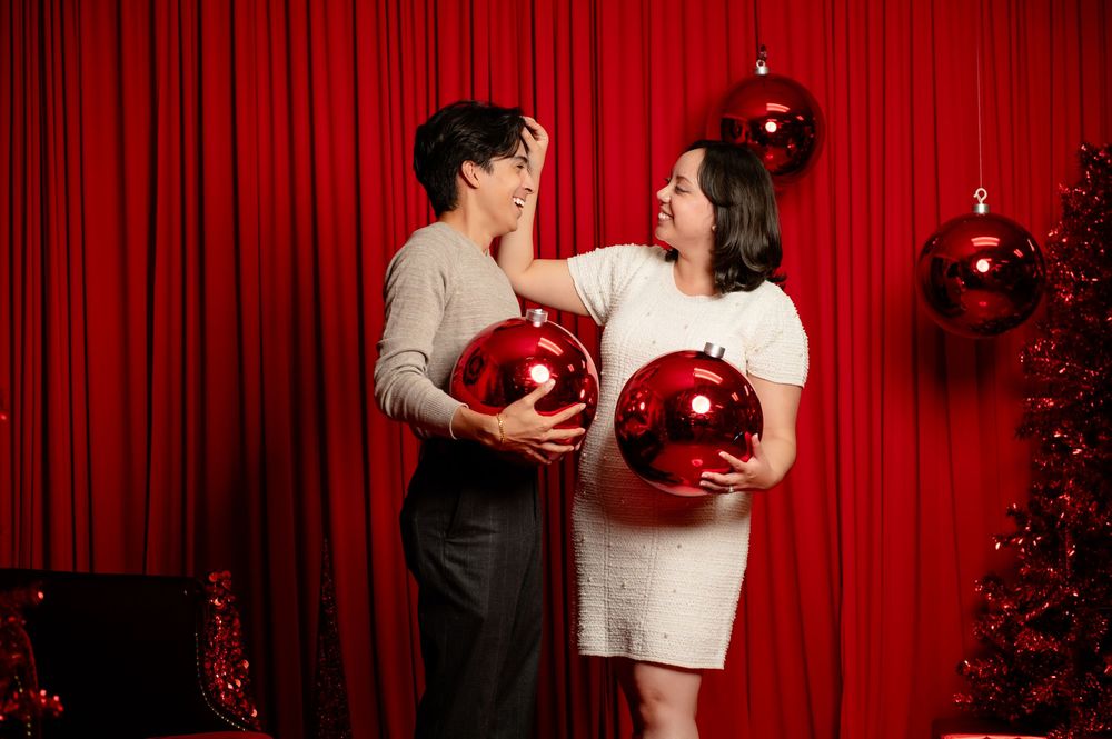 Two people laugh and hold large red Christmas ornaments in front of a red curtain beside a decorated red holiday tree.