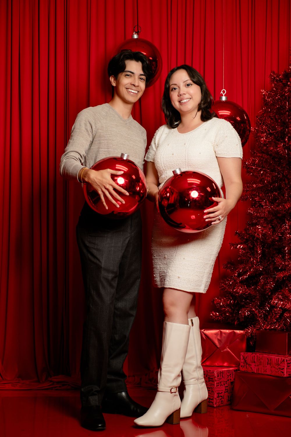 Smiling couple holding giant red Christmas ornaments in front of a red curtain beside a decorated red holiday tree and gift boxes.