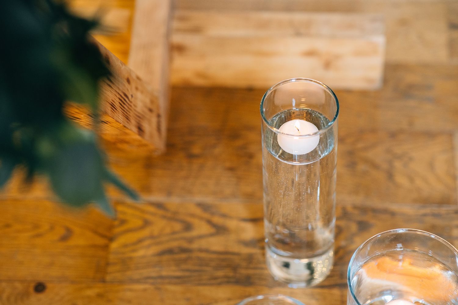 Floating white candle in a tall clear glass cylinder vase filled with water on a rustic wooden table beside greenery and a woven basket.
