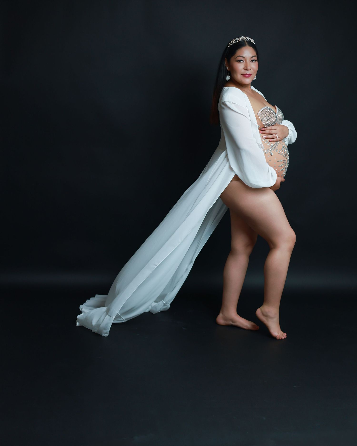 Pregnant woman in elegant white flowing robe posing for maternity photoshoot against dark background.