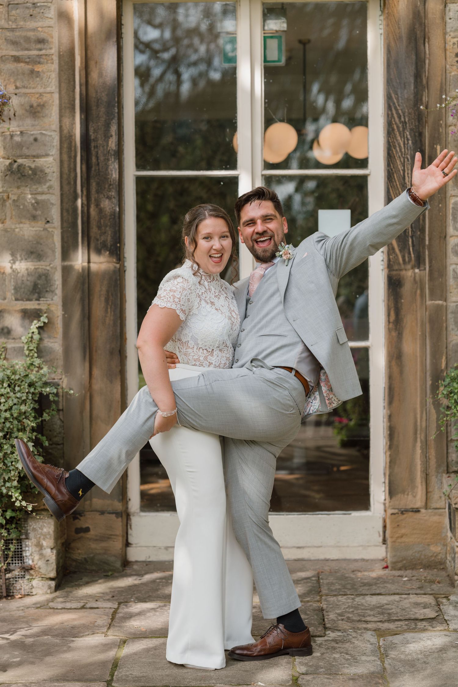 A joyful bride and groom celebrate outside a stone building, with the groom lifting his leg and waving in a playful pose.