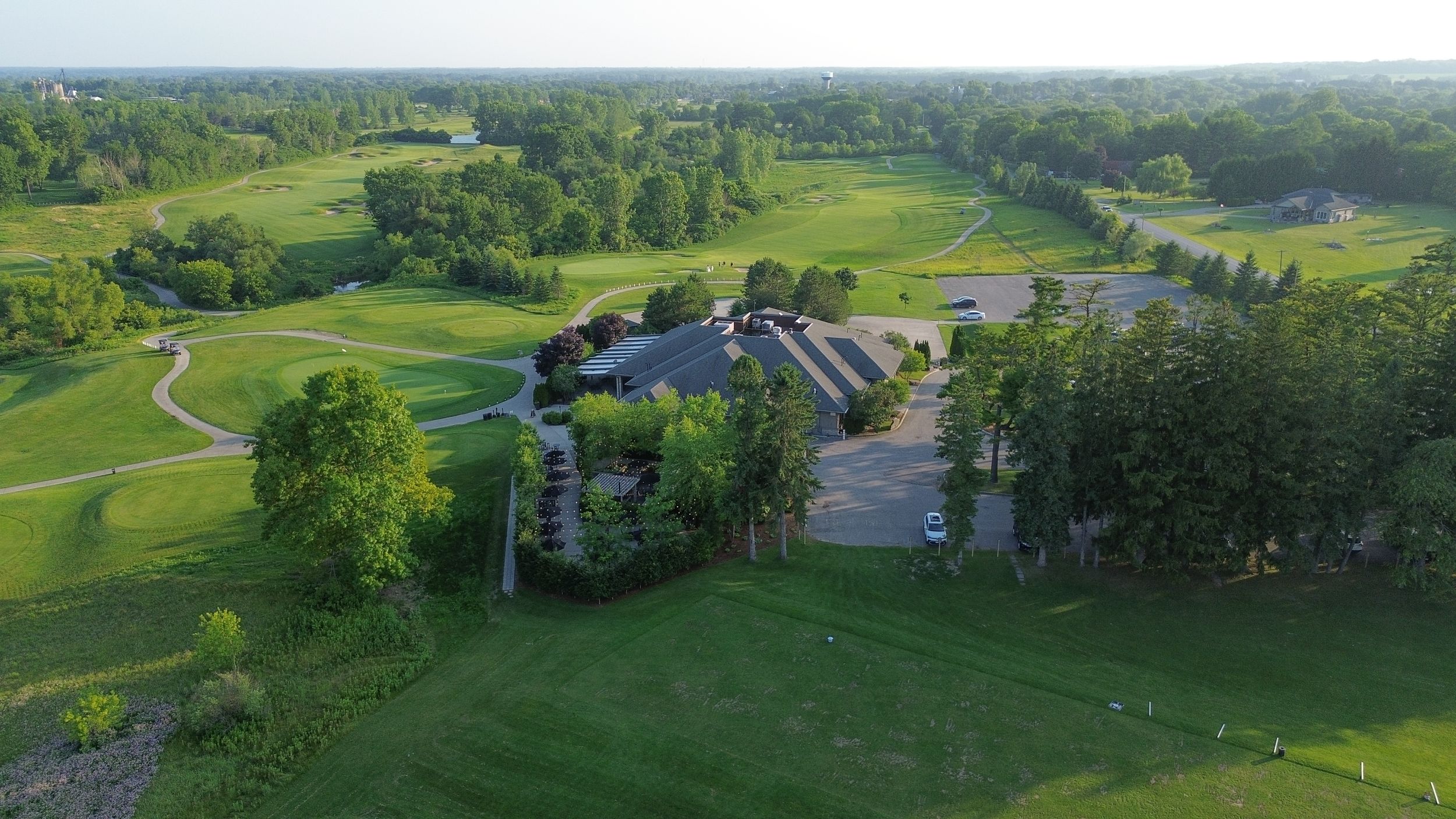 Aerial view of a lush green golf course with a clubhouse, parking lot, and surrounding trees on a sunny day.