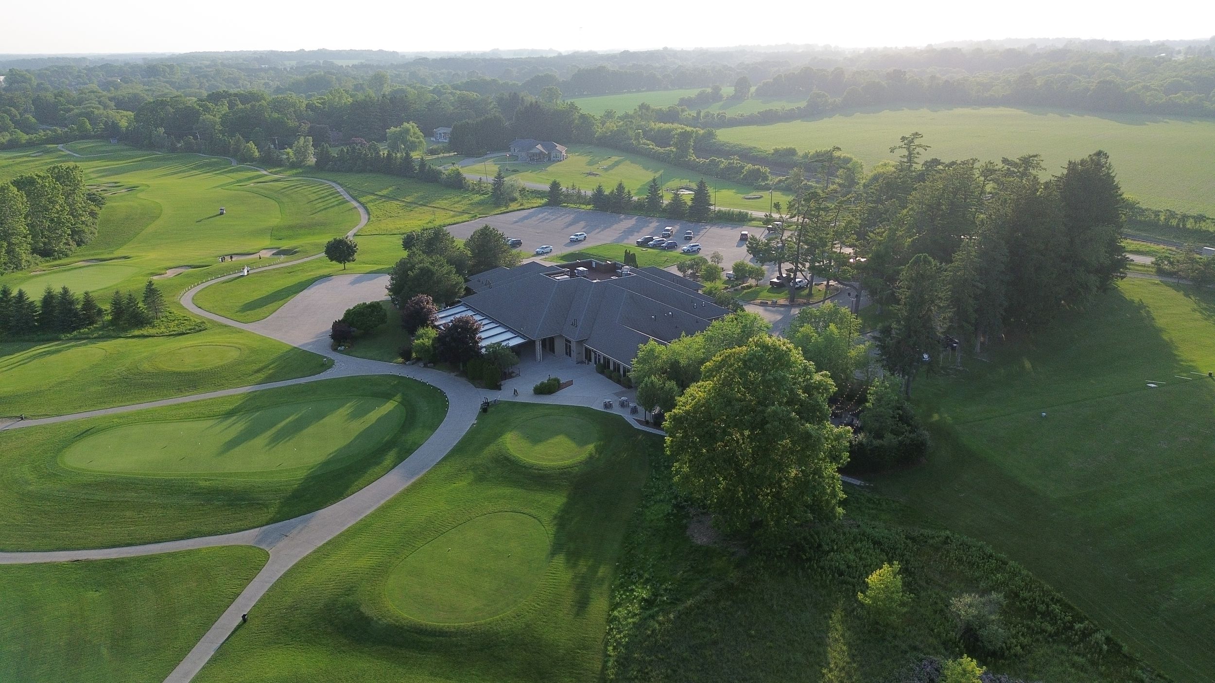 Aerial view of a lush green golf course clubhouse surrounded by trees and winding cart paths at sunset.