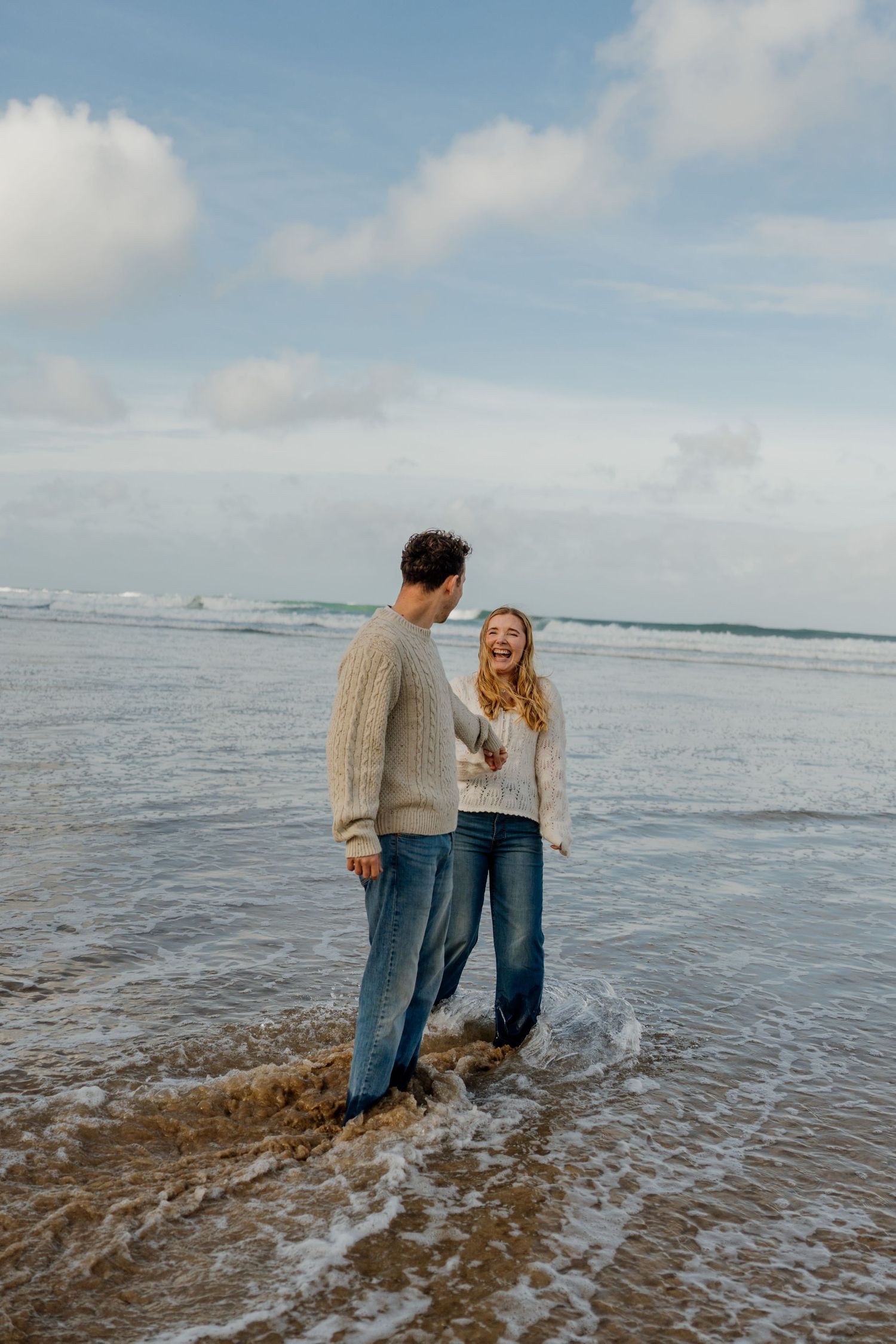 A happy couple in matching cream sweaters and jeans laughing together while standing in ocean waves at watergate bay.