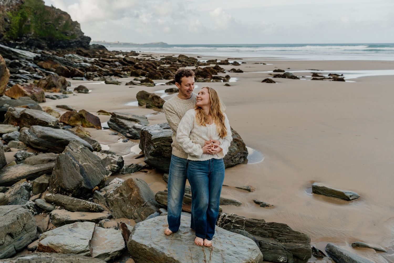 Couple embracing on rocky beach with sandy shore and ocean backdrop on a cloudy day at watergate bay in cornwall.