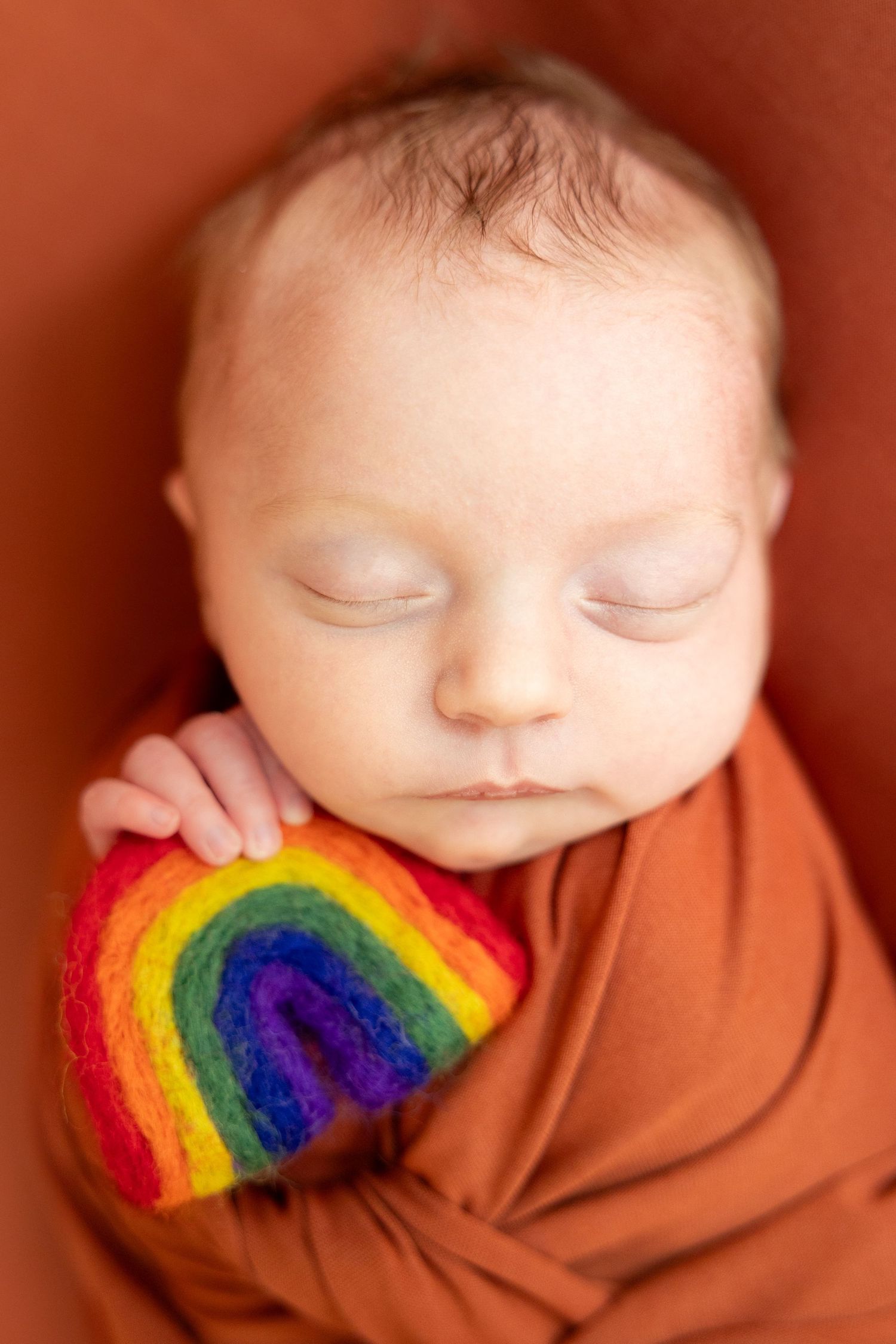 Sleeping newborn baby wrapped in rust orange swaddle holding a colorful felt rainbow toy.