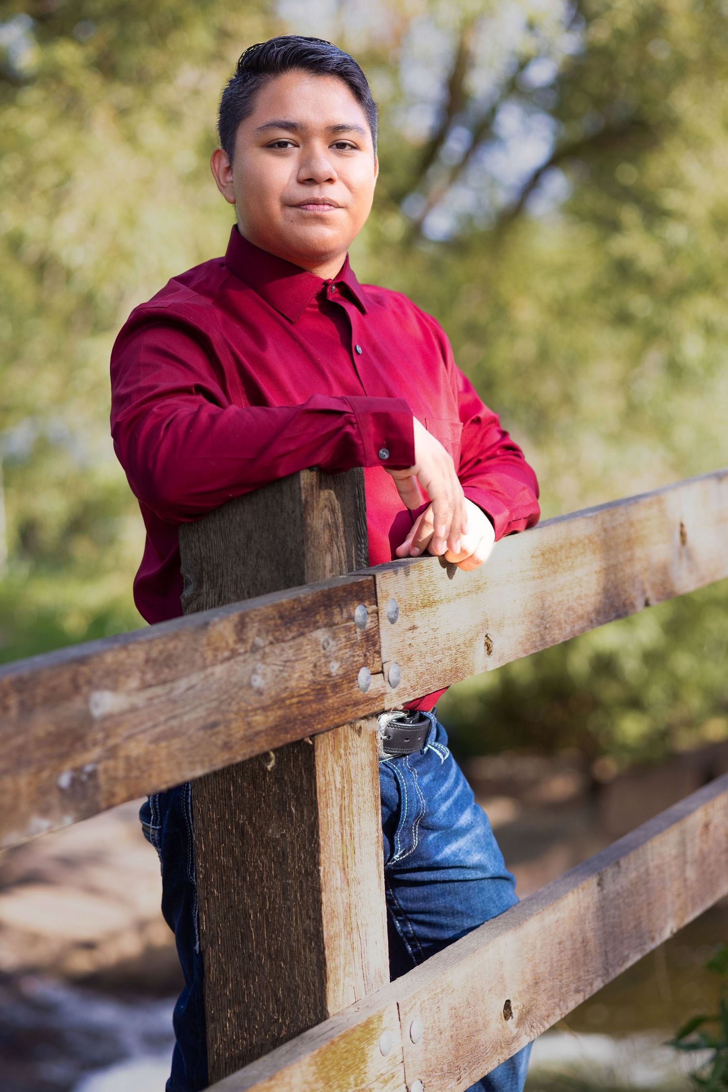 Young man in red shirt leaning on wooden fence outdoors, posing confidently with green trees in background.