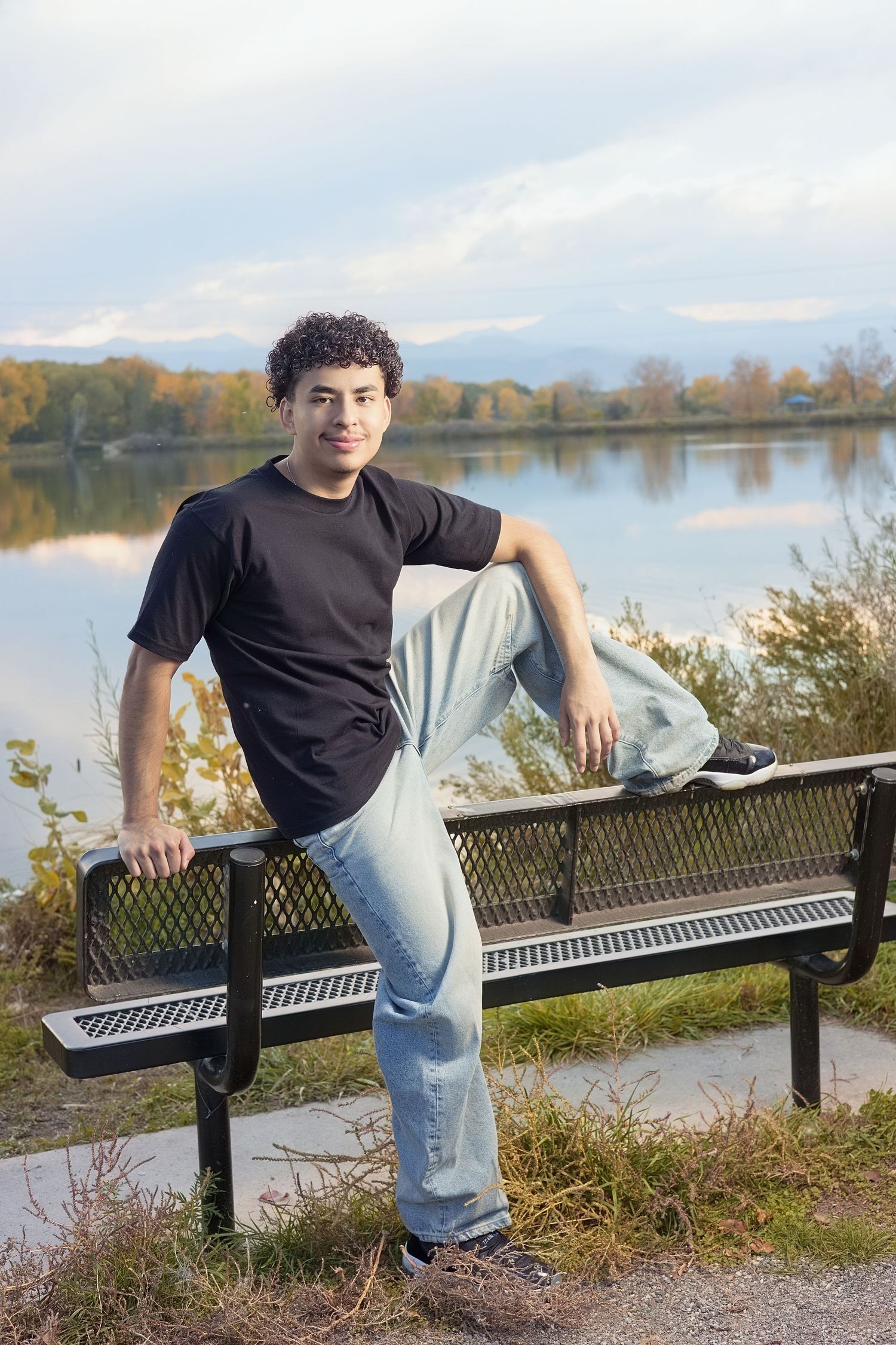 Young man in black t-shirt and jeans sitting on a park bench by a calm lake with autumn trees and mountains.