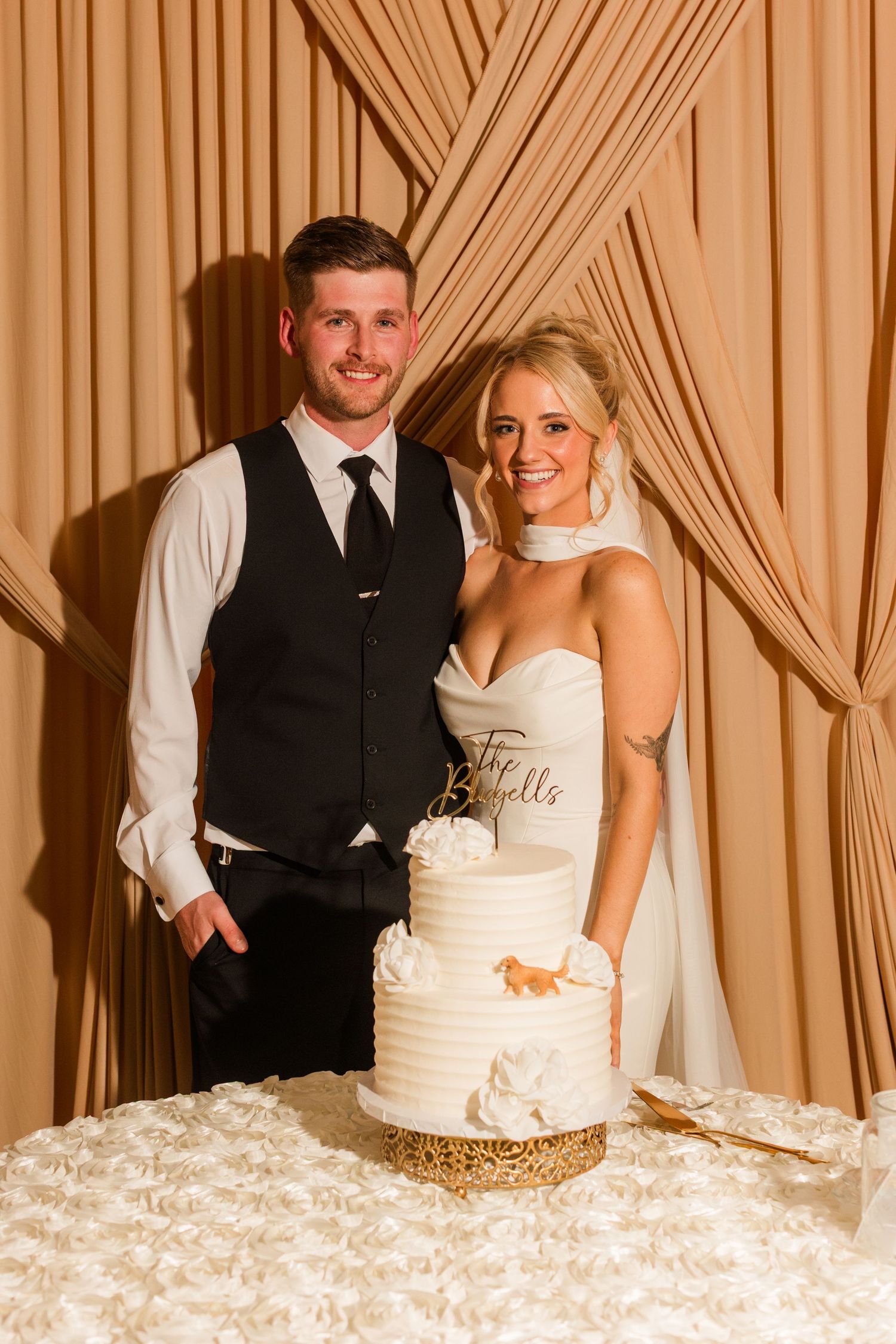 Bride and groom smiling together beside their white tiered wedding cake in front of elegant draped fabric backdrop.