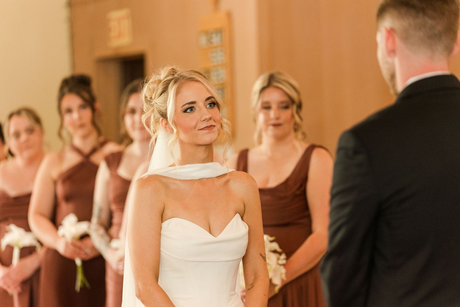 Bride in white strapless gown smiles tearfully at groom during wedding ceremony with bridesmaids in brown dresses behind her.