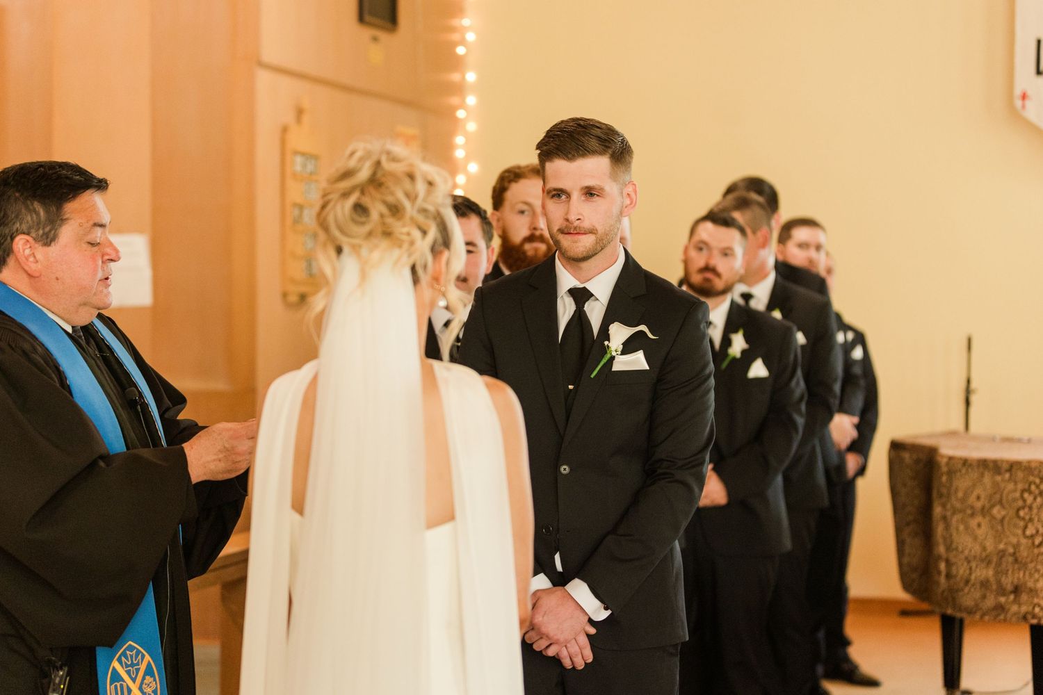 Groom in black suit holds bride's hands at church altar during wedding ceremony with officiant and groomsmen.