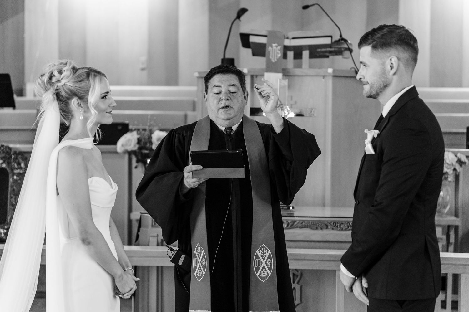 Black and white photo of a bride and groom exchanging vows with an officiant in a church wedding ceremony.