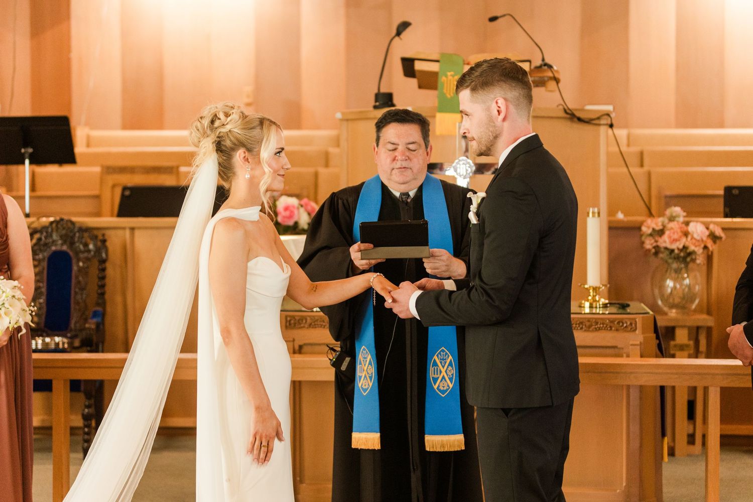 Bride and groom exchange rings during wedding ceremony at church altar with officiant in blue stole.