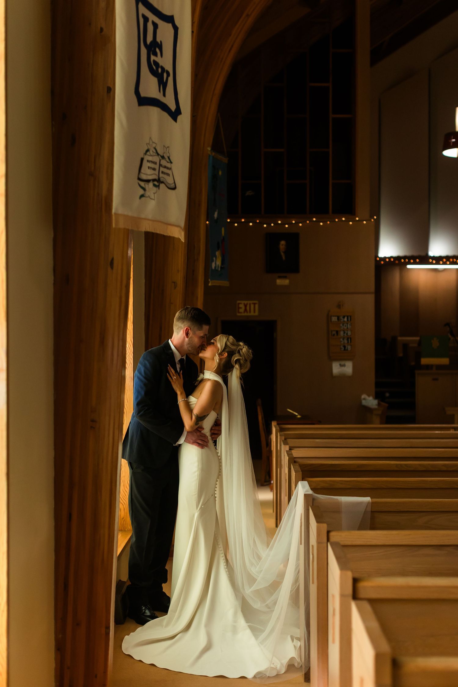 Bride and groom share a romantic kiss beside wooden columns inside a warmly lit church on their wedding day.