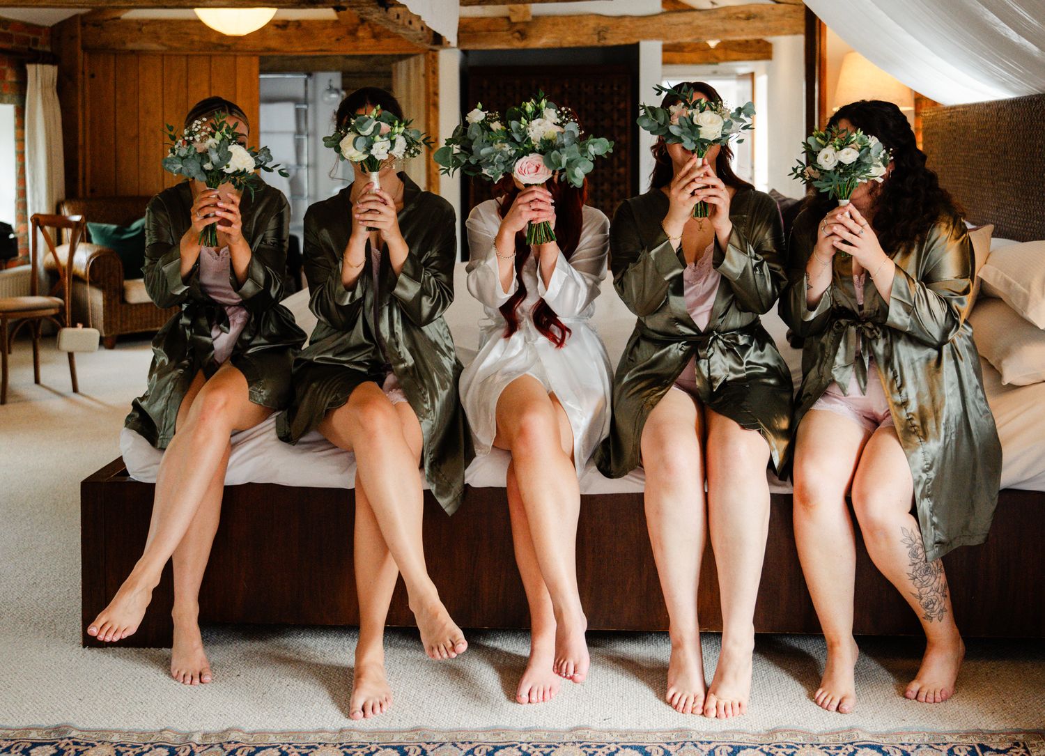 Five bridesmaids in satin robes sit on a bed hiding their faces behind floral bouquets during wedding preparations.