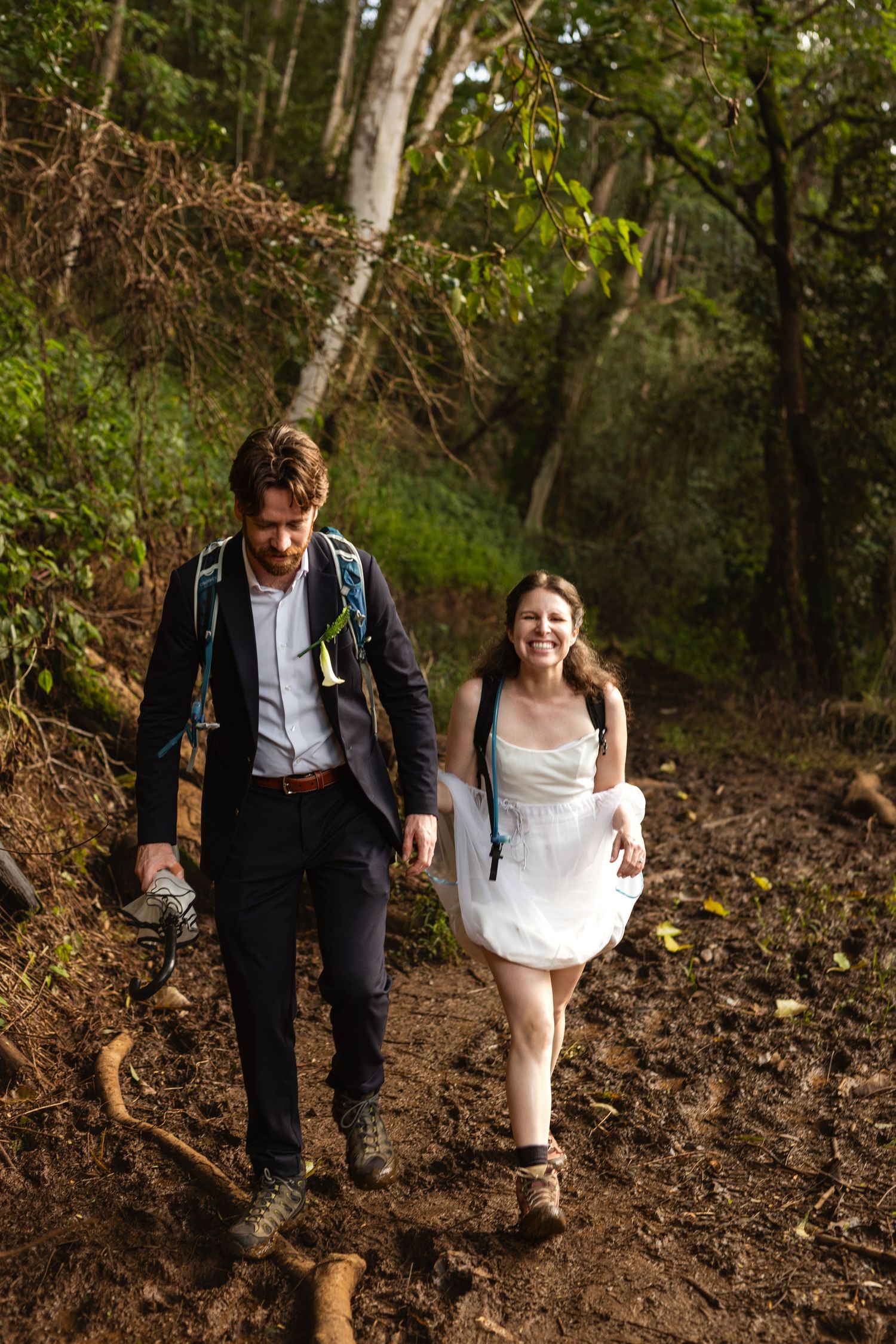 Smiling bride in a white dress and groom in a dark suit with backpacks hike a muddy forest trail during an adventure elopement.
