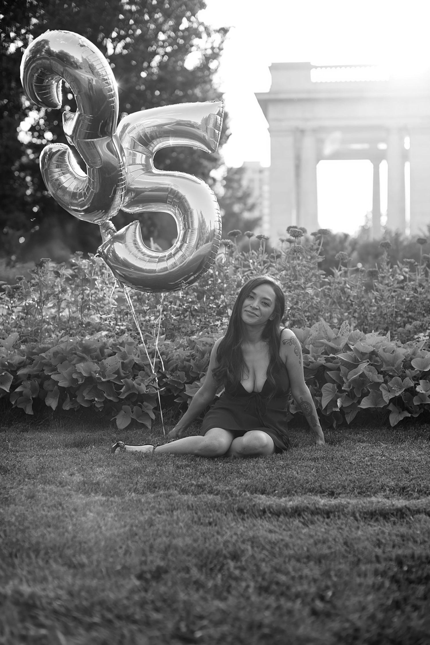 Black and white photo of a woman sitting on grass beside silver number 35 balloons, with garden shrubs and a columned monument behind her.