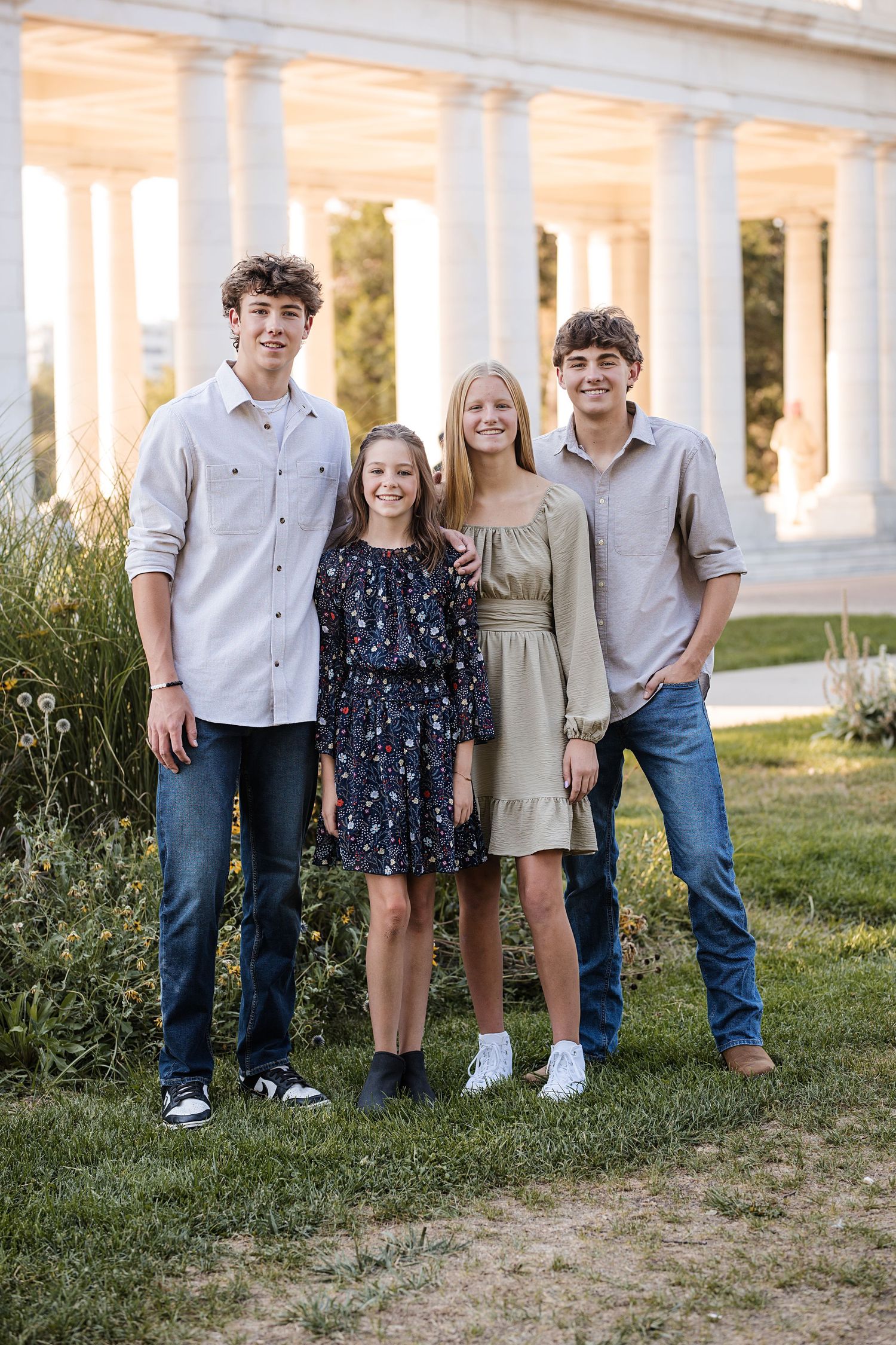 Four smiling teens and young adults pose together on a grassy lawn in front of a white columned colonnade during a family photo session.