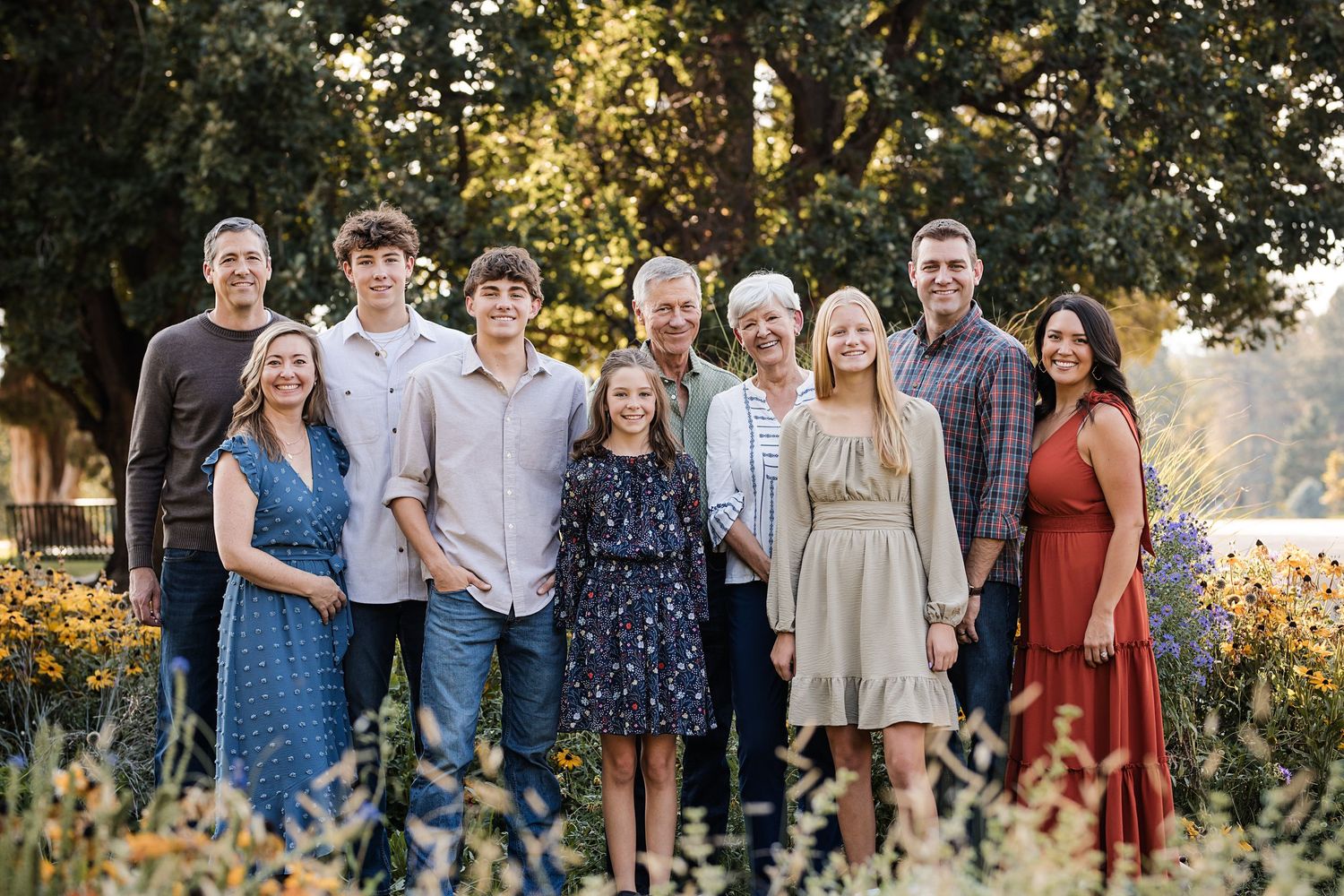 Multigenerational family of eleven posing together outdoors in a sunny garden with colorful flowers and tall trees in the background.