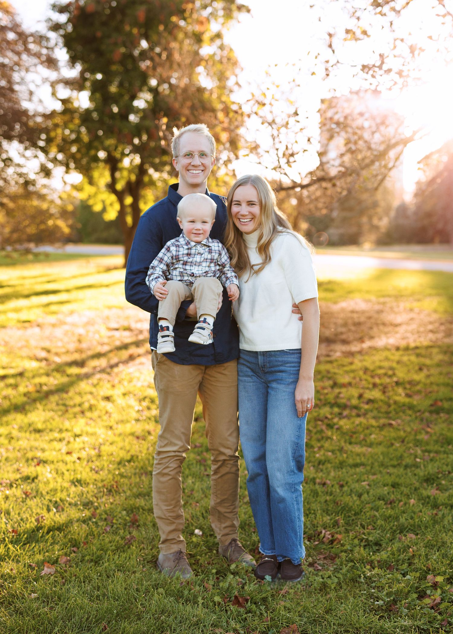 Smiling family of three with a man holding a toddler beside a woman, posing on a sunlit grassy lawn beneath autumn trees.