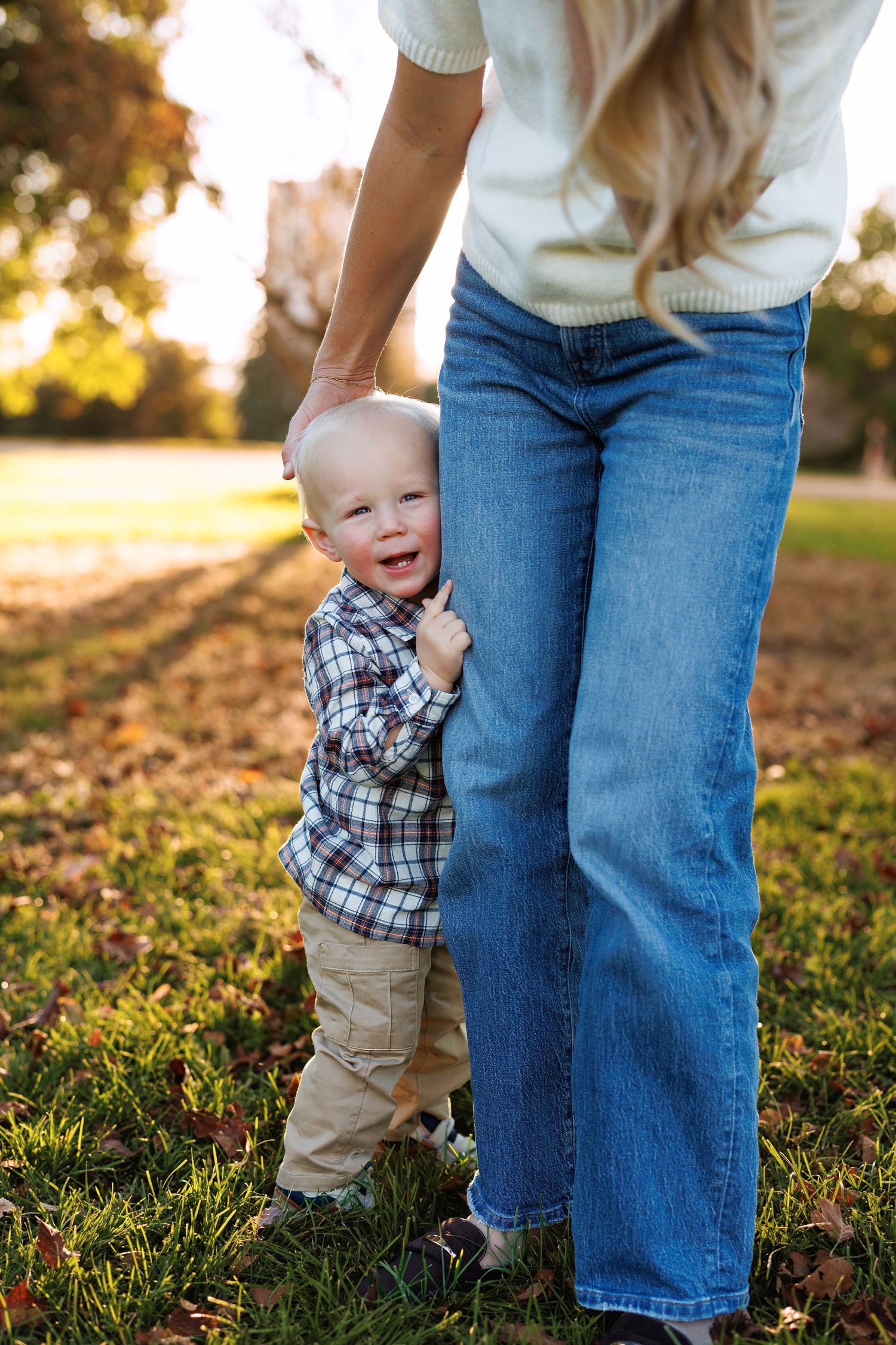 Smiling toddler in a plaid shirt hugs the leg of a woman in blue jeans on a sunny, leaf-covered lawn in autumn.