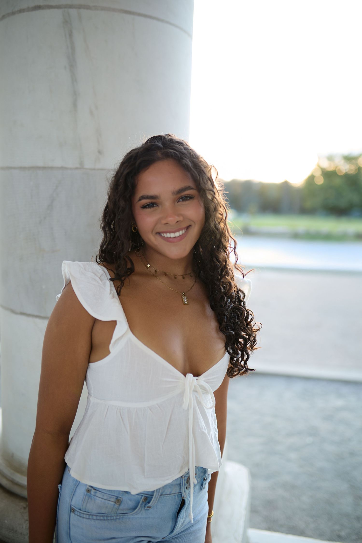 Smiling young woman with long curly dark hair in a white tie-front top and denim shorts leans against a marble column at sunset.