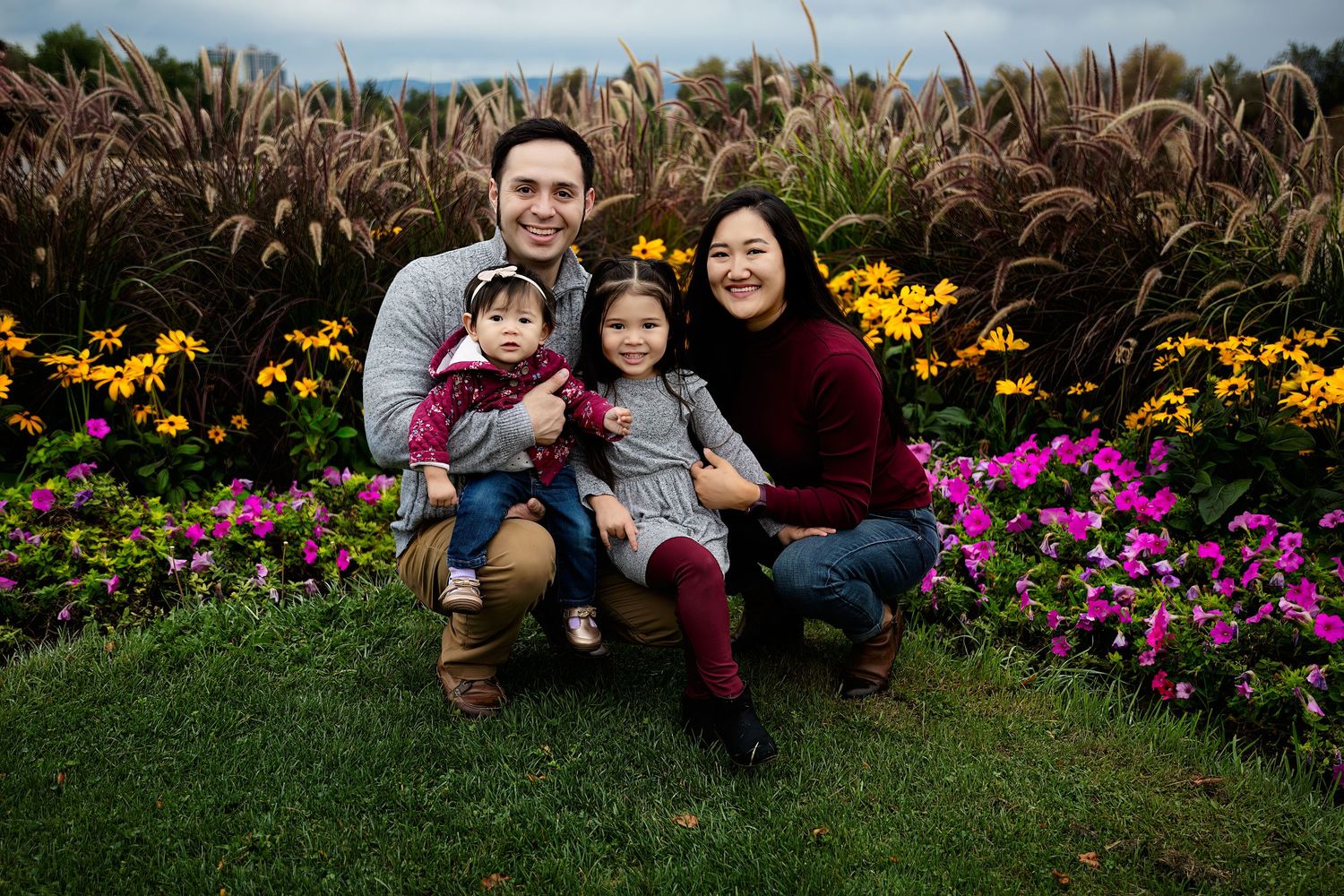 Smiling family of four posing together on a grassy lawn in front of colorful pink and orange flower beds and tall ornamental grasses.