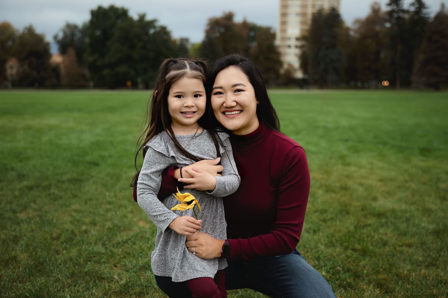 A smiling mother in a maroon sweater kneels on a grassy park lawn, hugging her young daughter in a gray cardigan, with trees behind them.