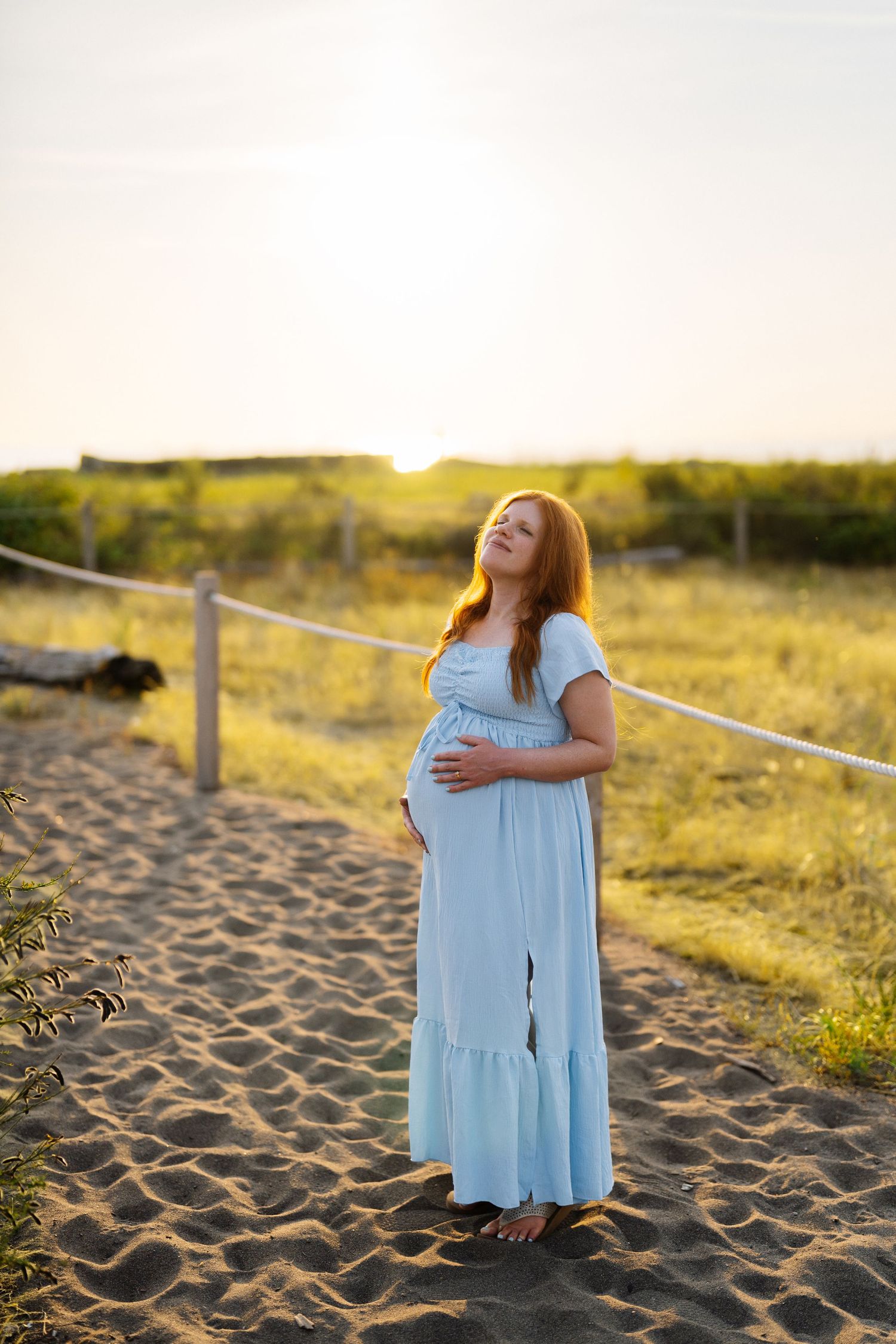 Pregnant woman in a light blue dress cradling her belly and gazing upward on a sandy beach path at golden hour sunset.