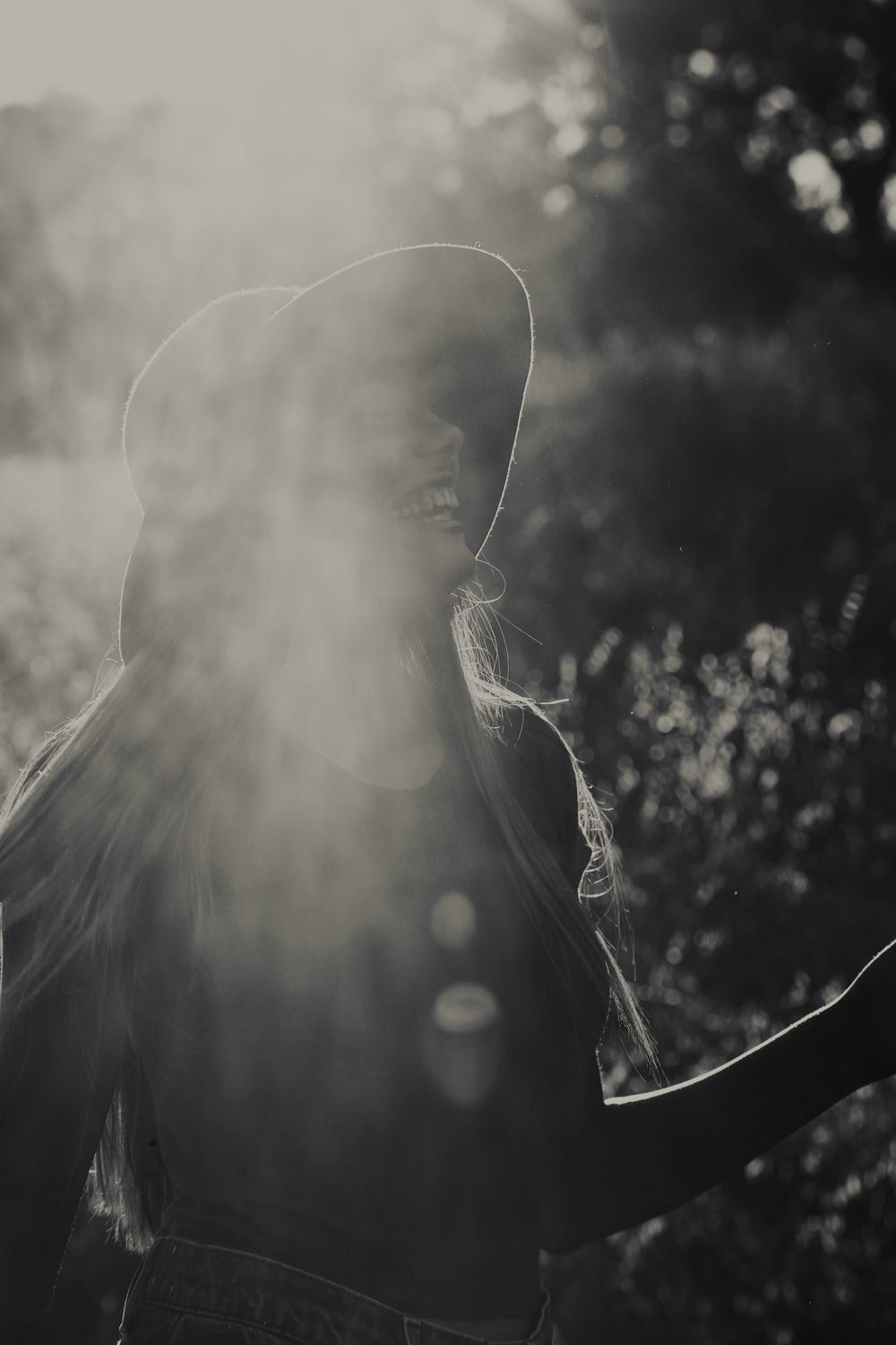 Black and white backlit image of a woman in a hat with arm outstretched, surrounded by dreamy sunlight haze and foliage.