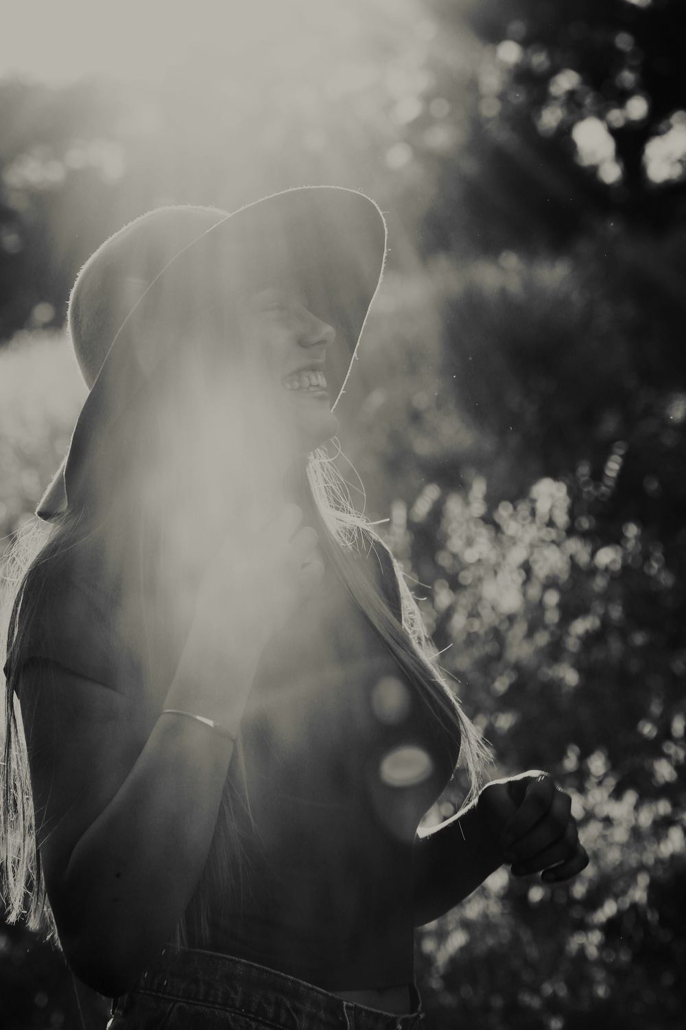 Monochrome photo of a laughing woman in a floppy hat standing outdoors, with soft sunlight haze and leafy bushes behind her.