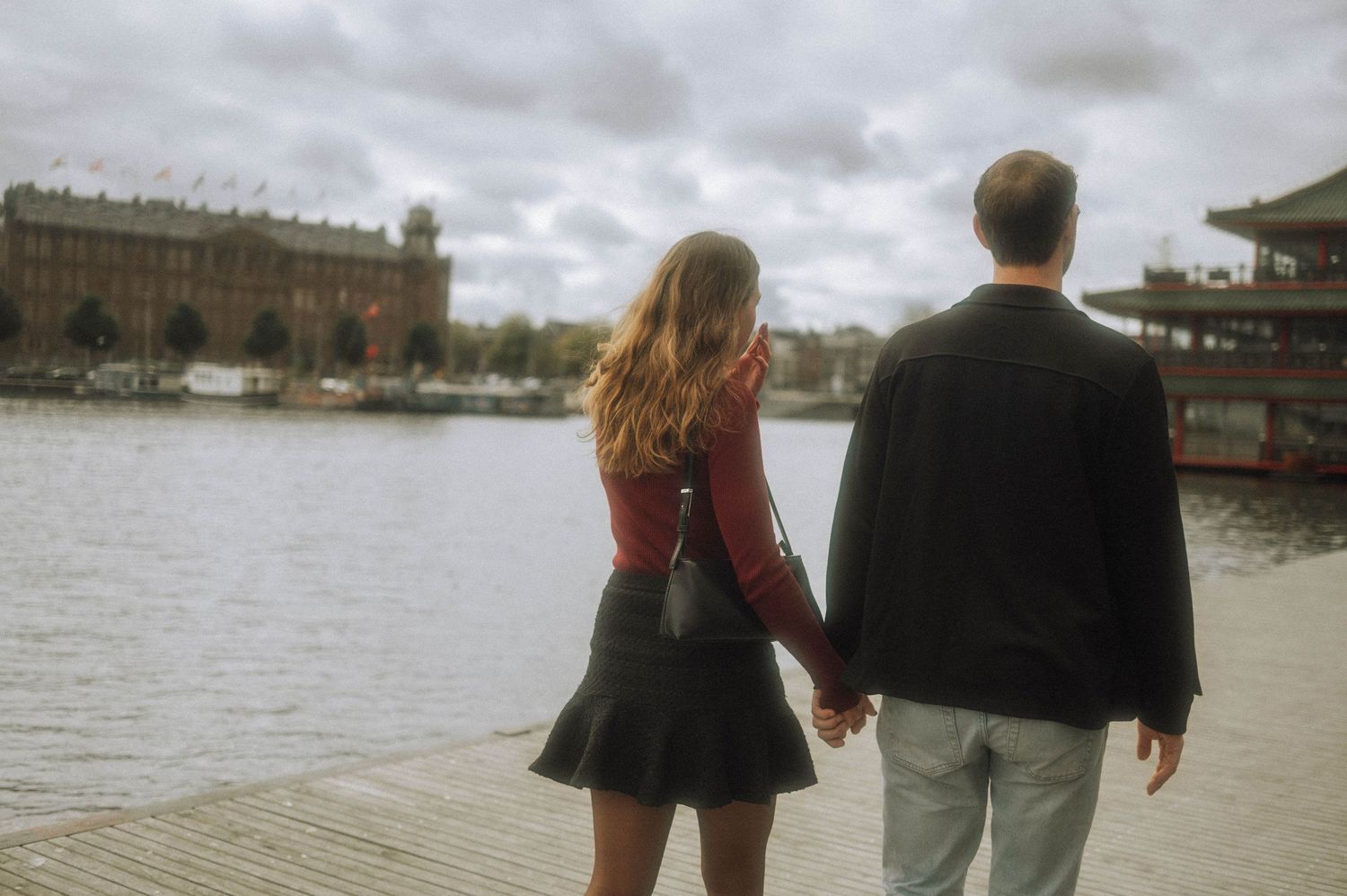 Couple holding hands while standing on a wooden waterfront pier, looking across calm water toward historic buildings in Amsterdam.