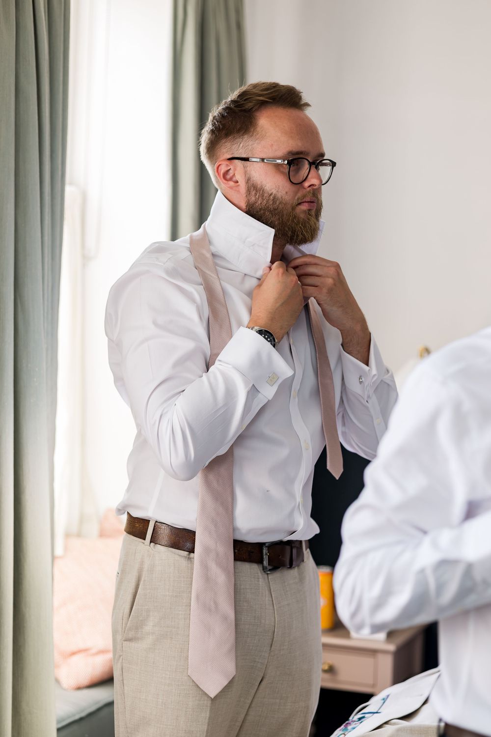Bearded groom in glasses adjusts his pink tie while getting ready for his wedding day.