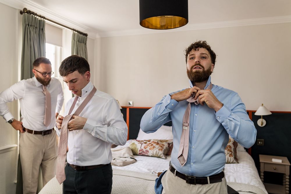 Three groomsmen adjusting their ties in a hotel room while preparing for a wedding ceremony.