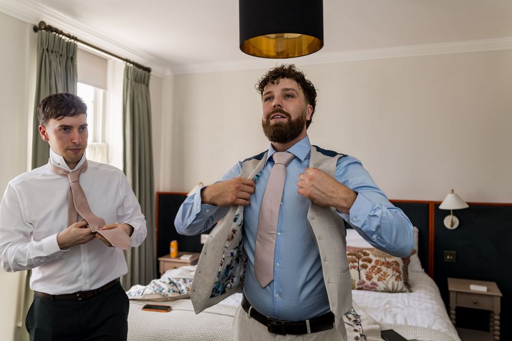 Two groomsmen in dress shirts and ties getting ready in a hotel room before a wedding ceremony.