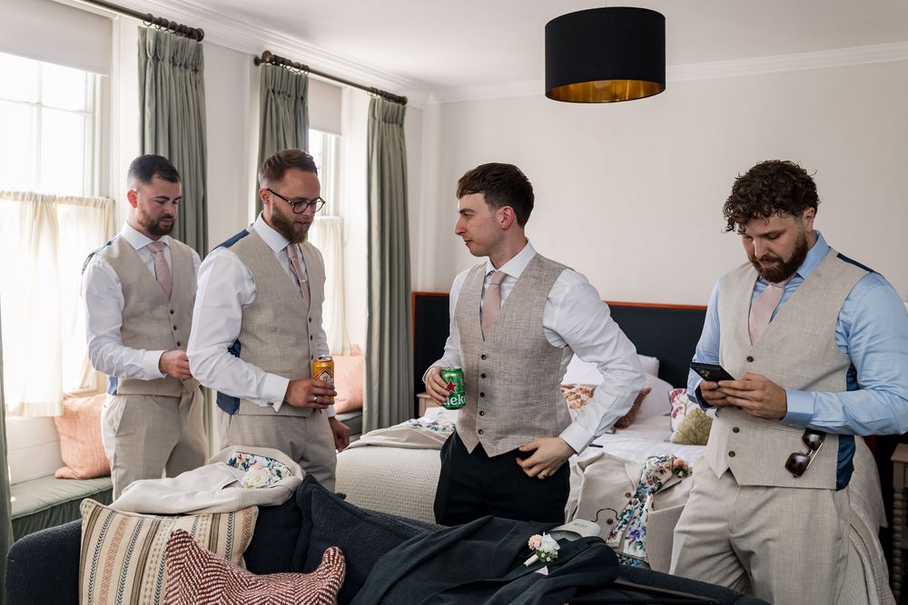 Four groomsmen in matching grey vests preparing for a wedding in a hotel room.
