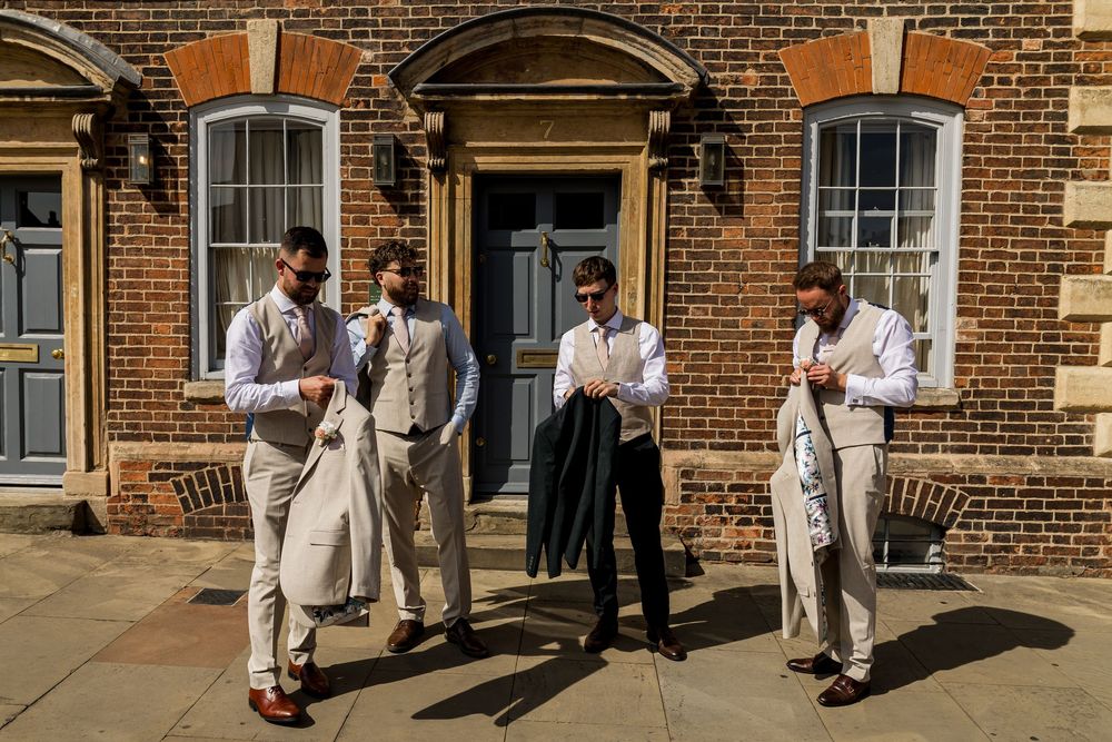 Four groomsmen in linen suits adjusting their attire outside a brick building on a sunny wedding day.