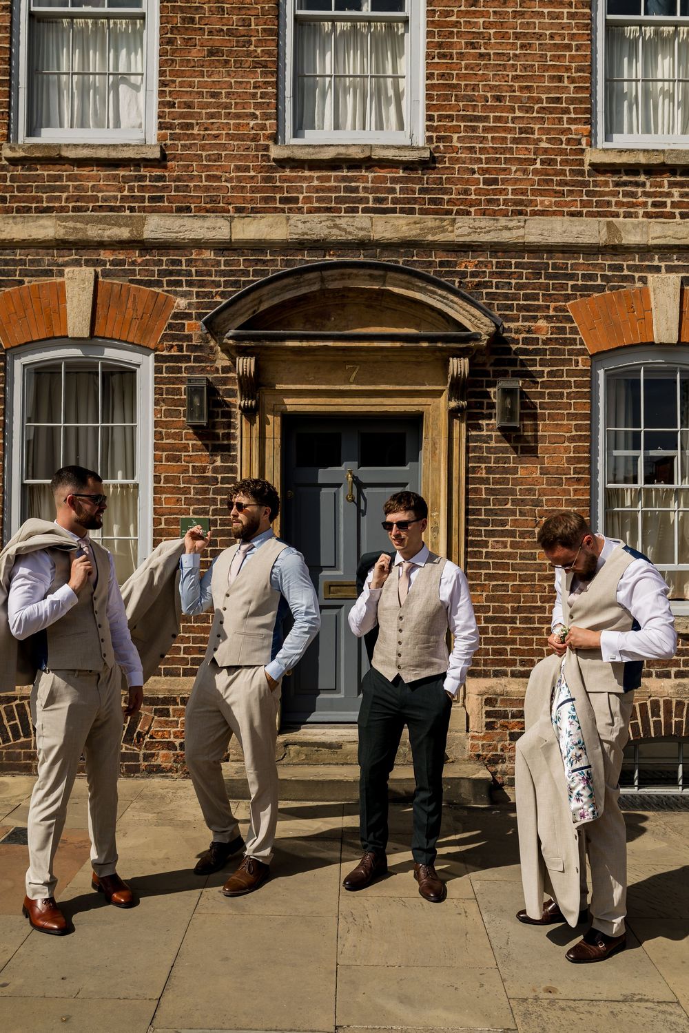 Four groomsmen in waistcoats and suits stand outside a Georgian brick building on a sunny wedding day.