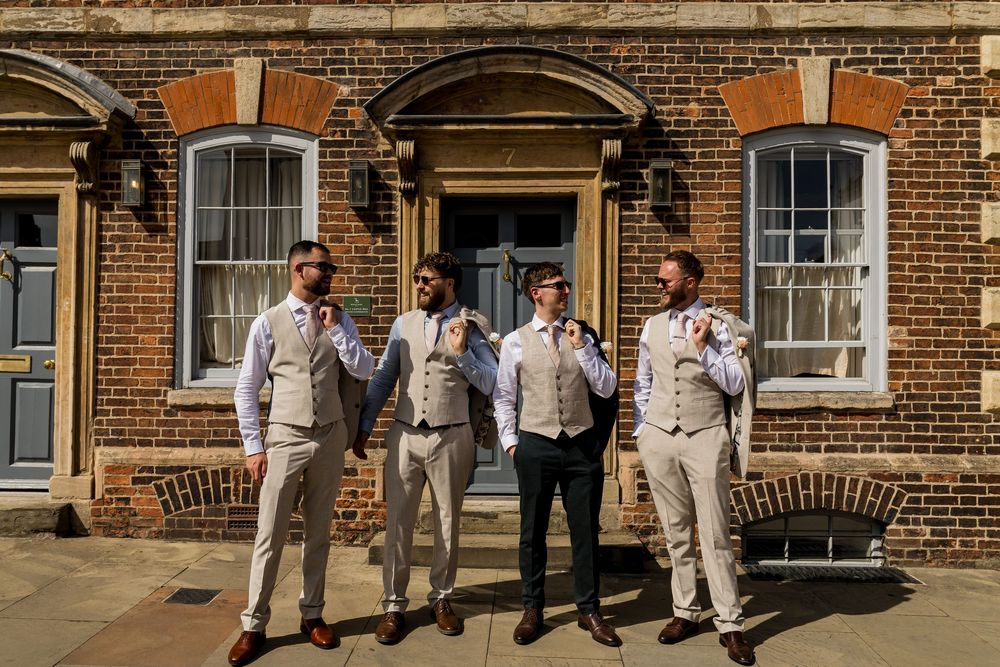 Four groomsmen in tan and black suits posing outside a brick building on a sunny wedding day.