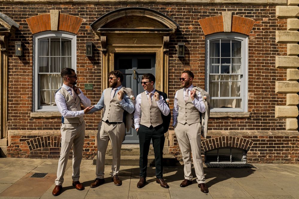 Four groomsmen in beige suits adjusting ties outside a brick building on a sunny wedding day.