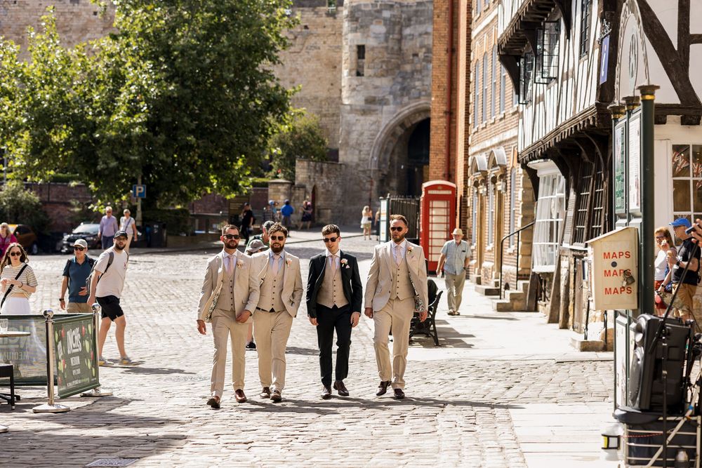 Four groomsmen in matching beige suits walking confidently through a historic cobblestone street.