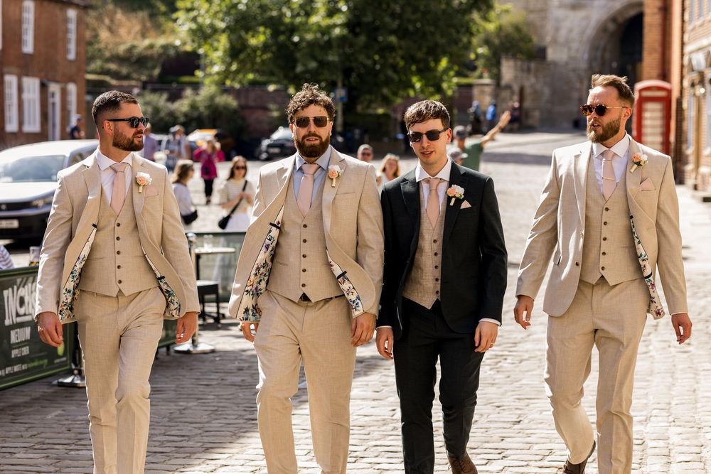 Four groomsmen in beige suits walking confidently on cobblestone street on a sunny wedding day.