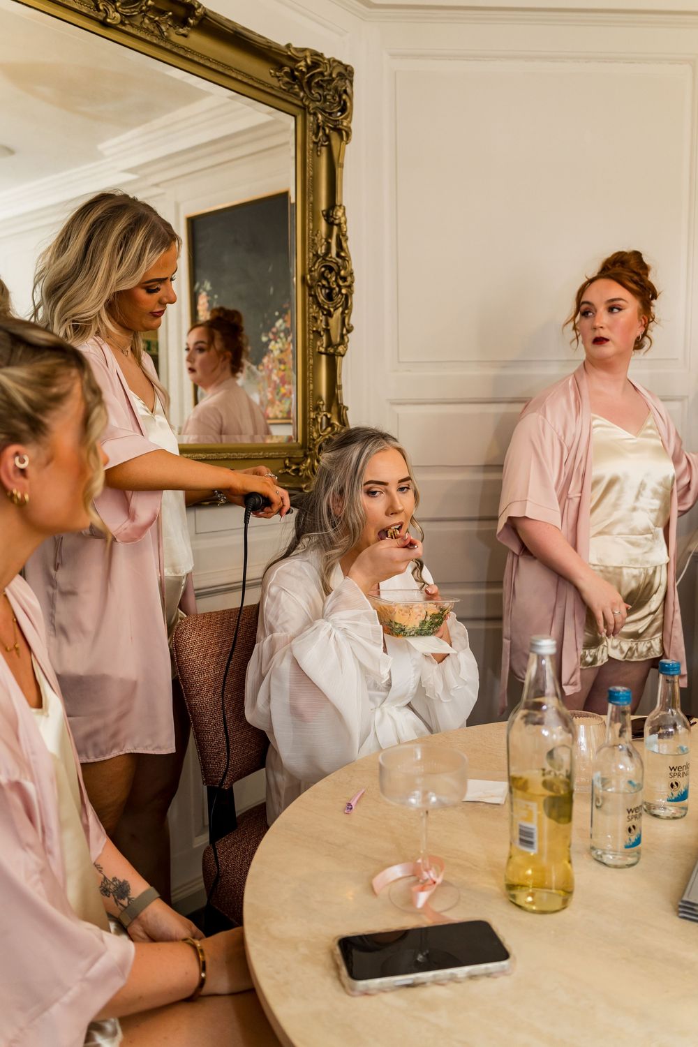 Bridesmaids in pink robes getting ready together, sipping drinks near a mirror in an elegant room.
